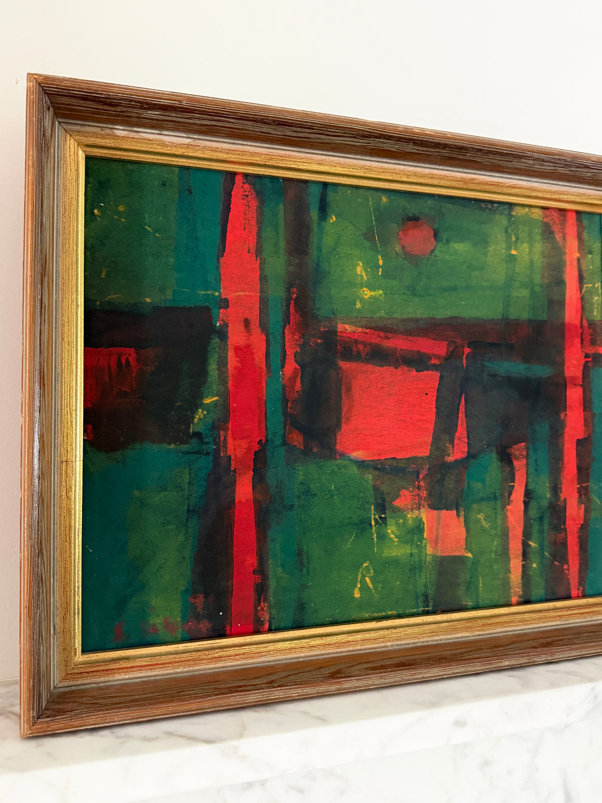 Framed abstract painting with bold green, red, and black brushstrokes, housed in a rustic brown wooden frame with a gold inner border, resting on a white marble surface against a white wall.